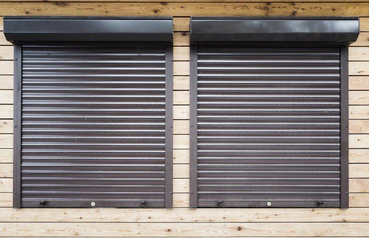 Dos ventanas con persianas oscuras bajadas y cajón exterior visto en una fachada revestida de madera